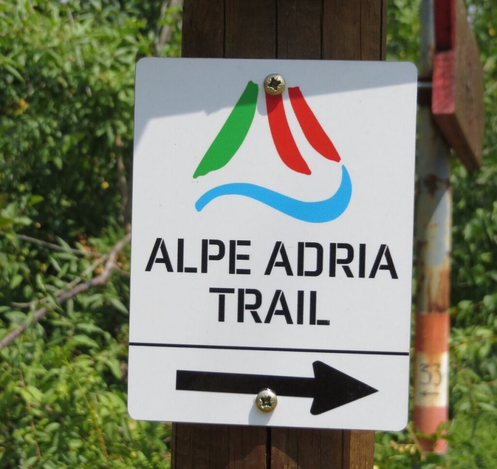 Cartello bianco dell'Alpe Adria Trail con il logo e una freccia, fissato a un palo